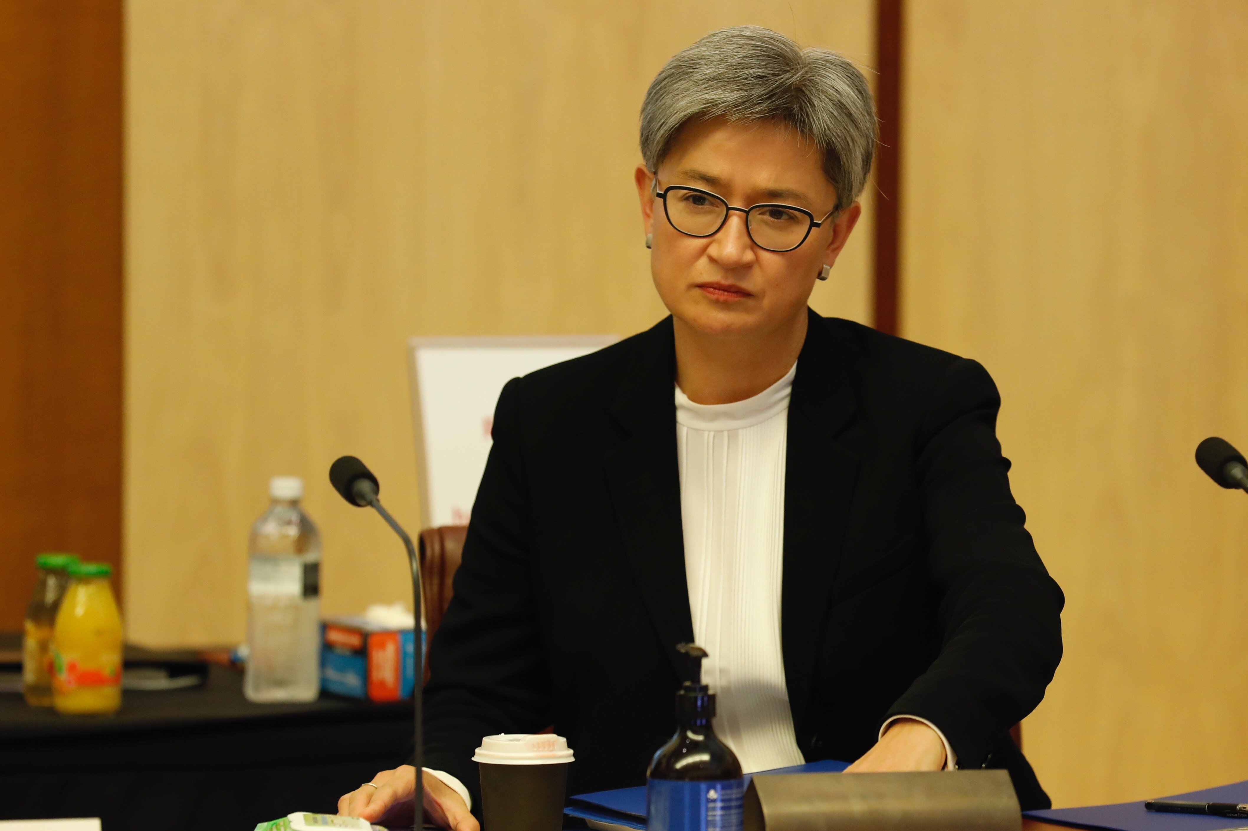 A woman with short grey hair and glasses wearing a blazer and white shirt sitting at a desk behind a microphone