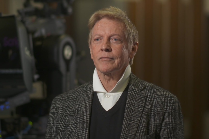 A man in a blazer, sweater, and a shirt with a turned-up collar looking serious.