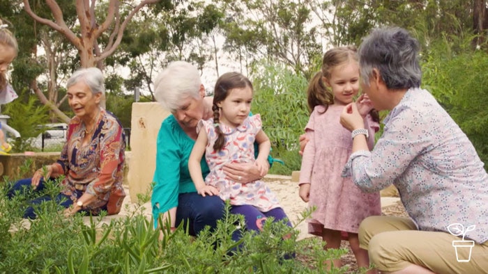 Elderly women sitting on the ground in a garden with two young girls (one girl giving a lady something to eat)