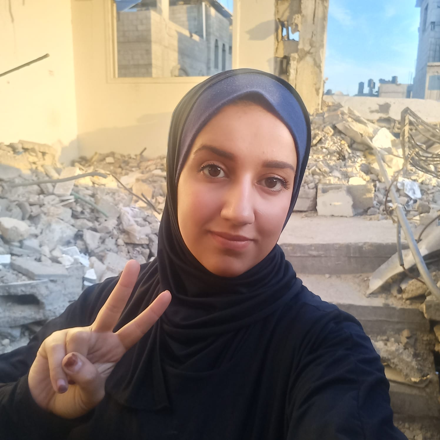 A Palestinian woman in a black headdress making a peace symbol with her right hand standing in front of mounds of rubble