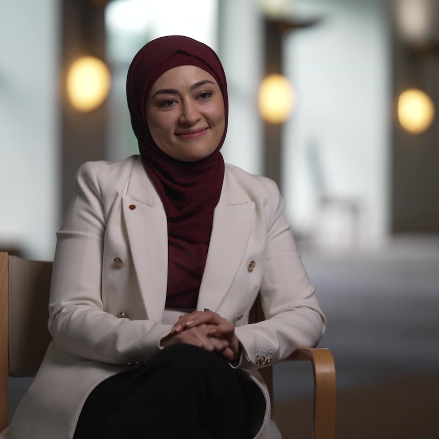 Fatima Payman wearing a maroon hijab and white blazer sits in a hall with the background out of focus