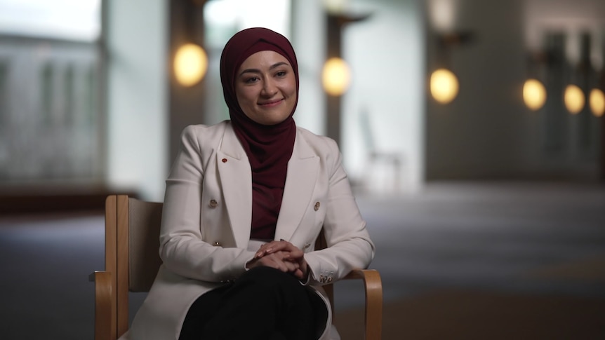 Fatima Payman wearing a maroon hijab and white blazer sits in a hall with the background out of focus