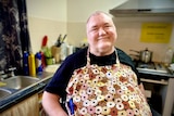 A man with short, grey hair wearing a black T-shirt and an apron with doughnuts on its sits in a wheelchair in his kitchen