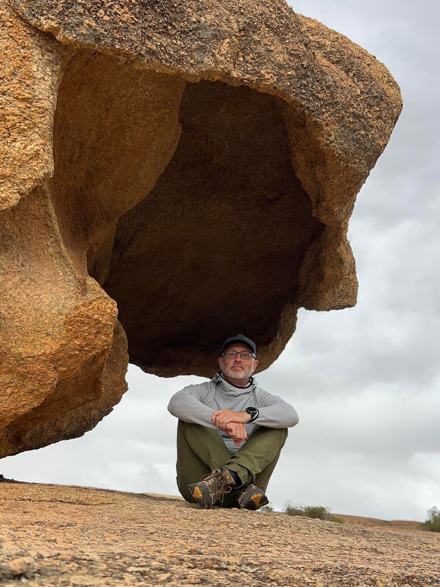 A man with a grey beard and glasses wearing hiking gear sits cross legged underneath a rock formation. 