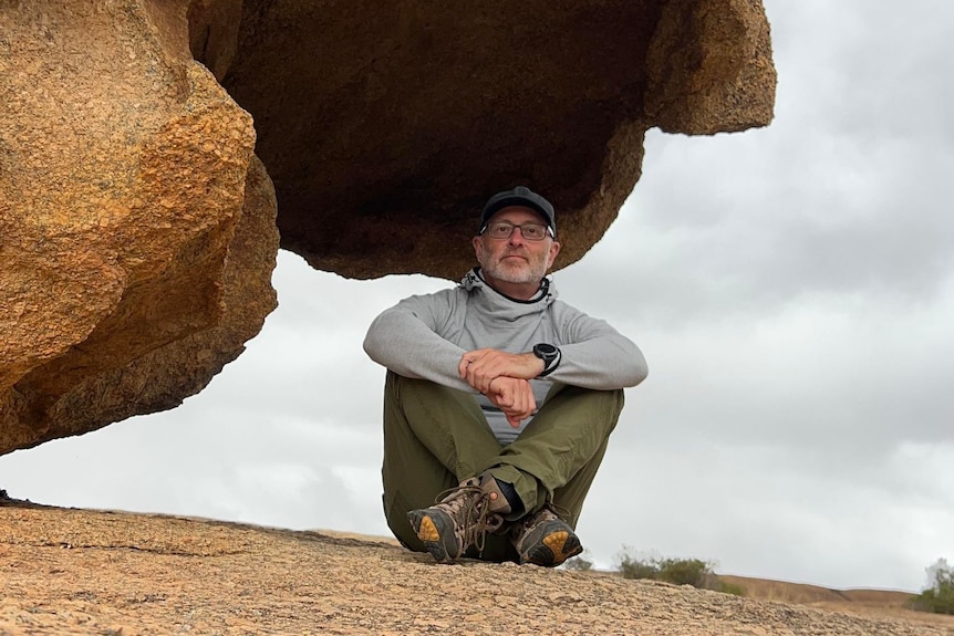 A man with a grey beard and glasses wearing hiking gear sits cross legged underneath a rock formation. 