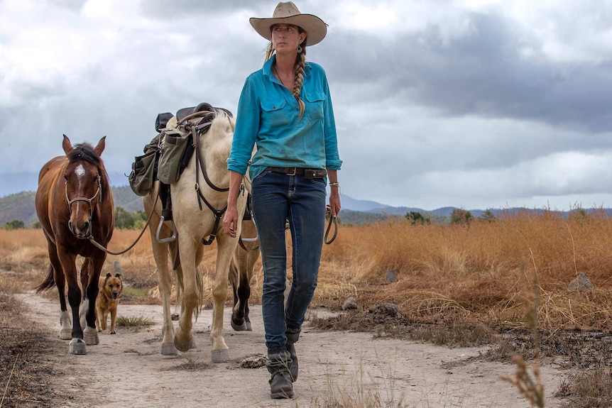 A woman walks in front of her three horses and dog.