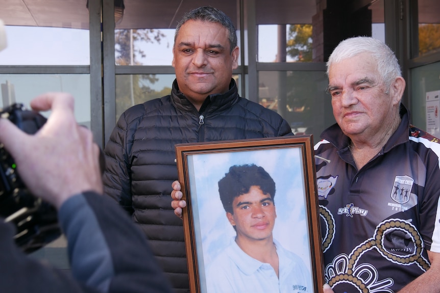 Two men hold a picture of a teenage boy who was found dead decades ago.