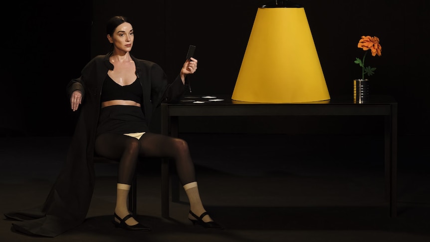 Annie Clark AKA St Vincent sitting in black clothing infront of a blank canvas with a lampshade and plant on a desk.