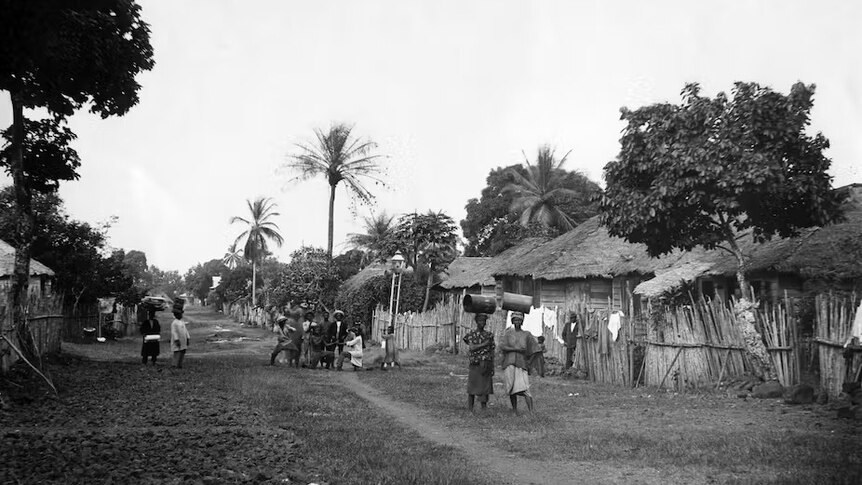 B&W pic of an African village. Thatched huts on right, palm trees & local women carrying containers on their heads