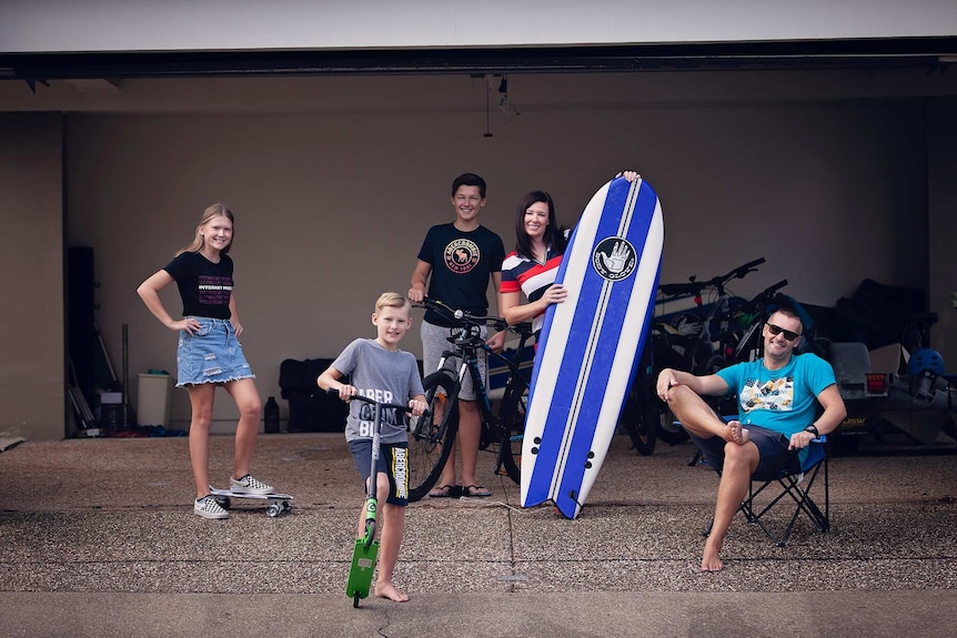 Man sitting on chair woman holding a surfboard with boy on bike, boy on scooter and girl on skateboard in a house garage