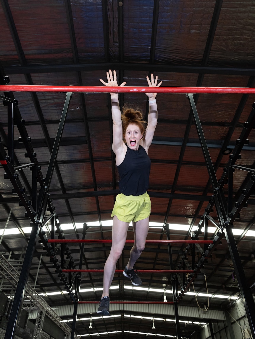 A woman leaps through the air to grab a red horizontal bar, grinning widely