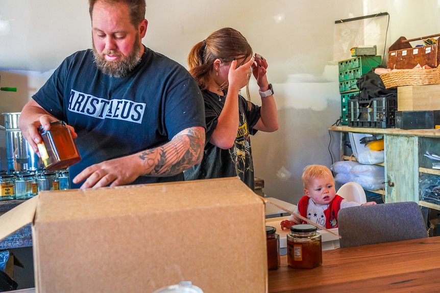 A bearded man with a tattoo packs honey jars into a large box on a table.