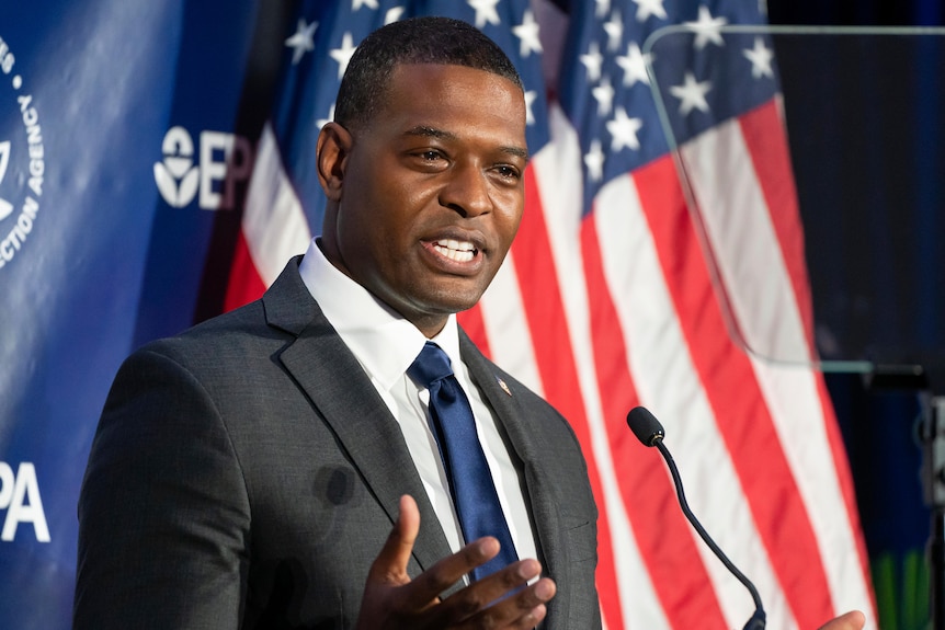 Un hombre negro de mediana edad con un traje gris habla por un micrófono frente a una bandera estadounidense y un fondo azul de la EPA.