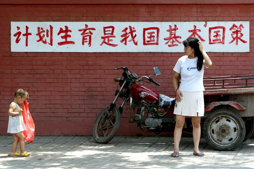A young Chinese mother watches her child walk in front of a sign reading "birth control is a basic state policy of our country".