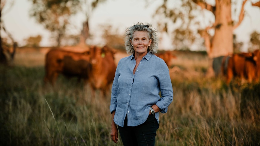 A middle aged woman with blonde hair and a blue collared shirt stands in a field with cattle in the background.