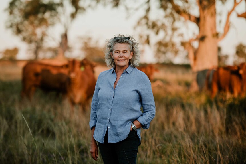 A middle aged woman with blonde hair and a blue collared shirt stands in a field with cattle in the background.