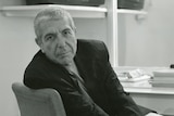 An older man sitting at a desk that is stacked with books. He is turned in his seat to look casually at the camera
