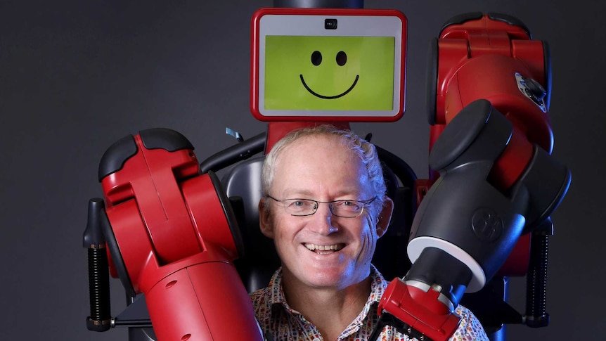 Baxter the robot wraps his arms around Toby Walsh, who is sitting on a chair in front of it
