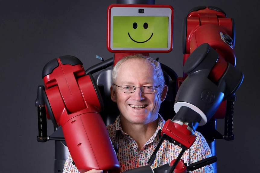 Baxter the robot wraps his arms around Toby Walsh, who is sitting on a chair in front of it