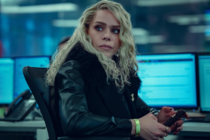 A blond woman in a black jacket sits in front of computer screens