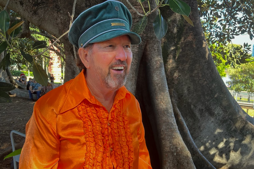 A man wearing green hat and bright orange shirt with ruffles smiles under a tree