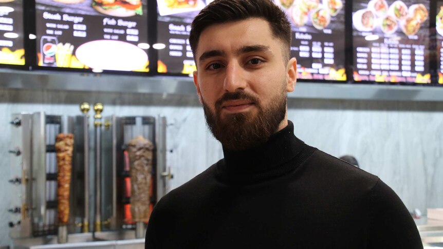 A head and shoulders shot of a man wearing a black turtleneck standing in a kebab shop looking at the camera.