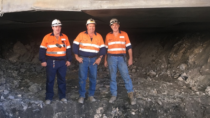 Three men in hard hats and high-vis standing in a line at a coal mine.