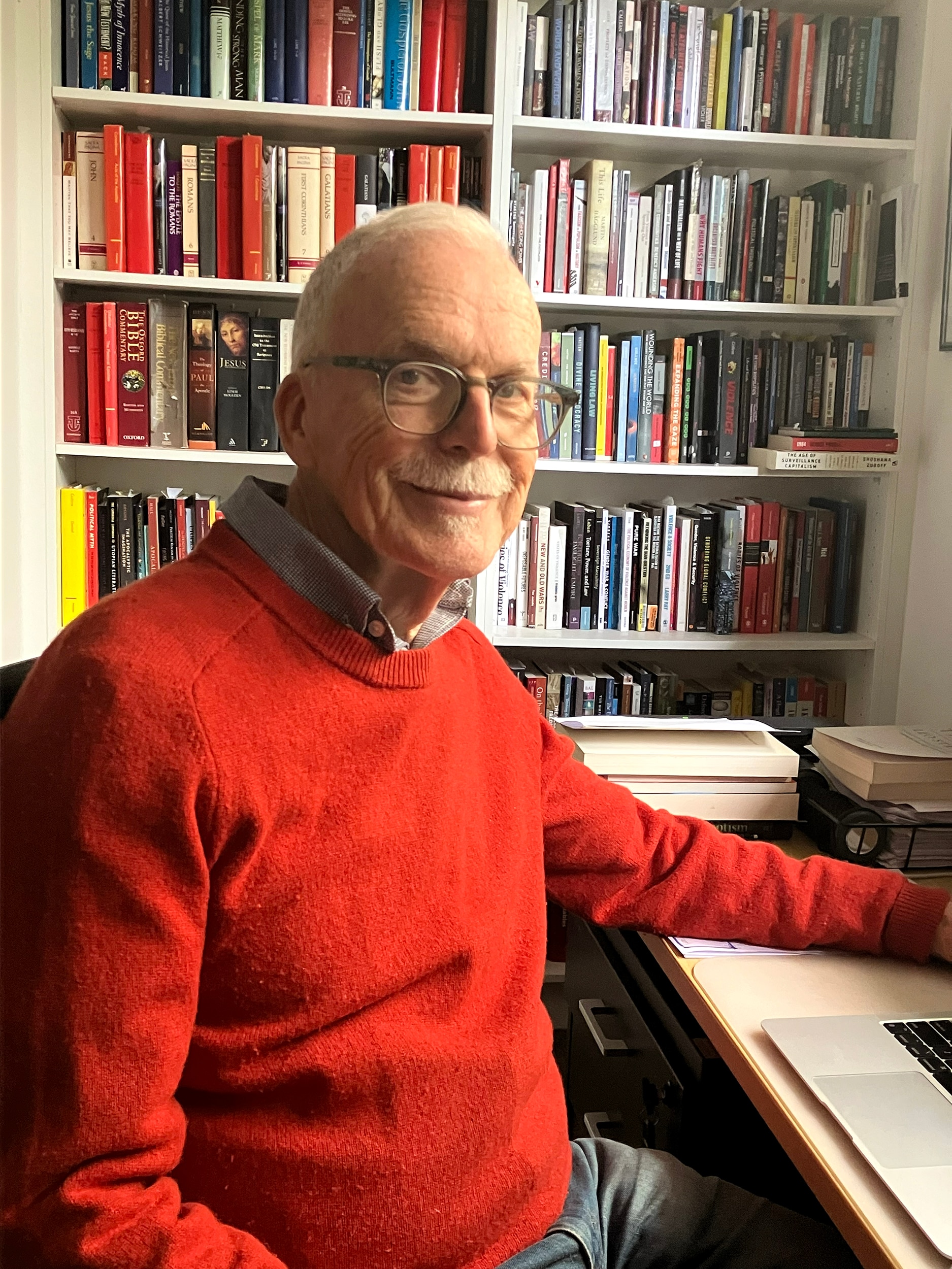 Man in red jumper sits at desk with bookshelf behind him.