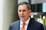 A close up of Jim speaking in a suit and orange tie outside a building during the day.