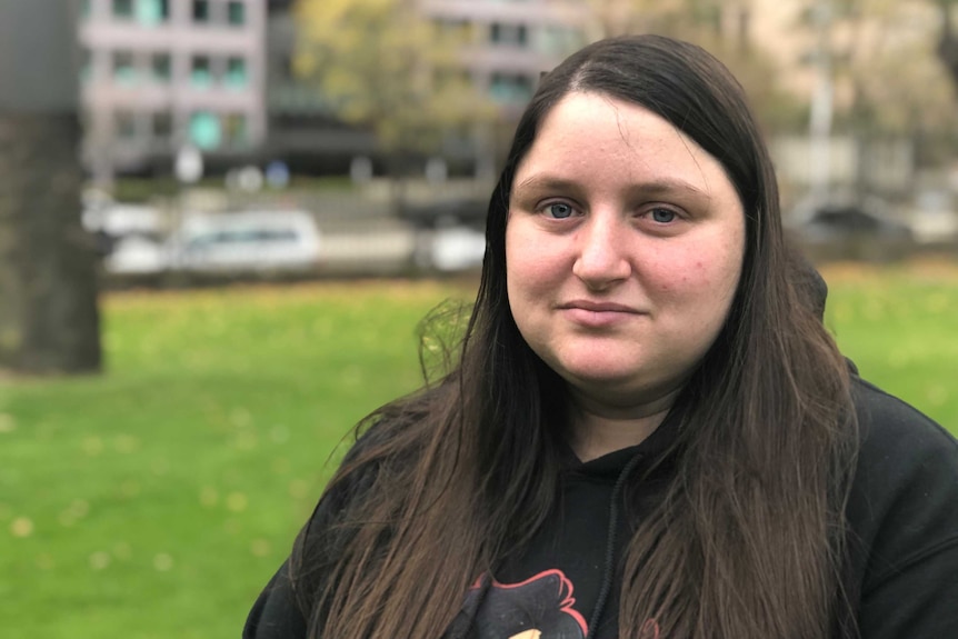 A young woman with long dark hair and a black t-shirt sits in a park.