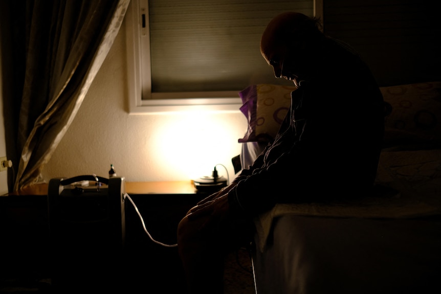 The silhouette of a man sitting on a bed with a tube running from a desk. 