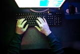 a person's hands can be seen on a black keyboard from above as a monitor glows on to the hands and desk and keyboard