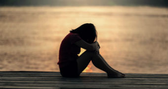 A woman silhouetted against the sunset on the water behind her rests her head on her knees.