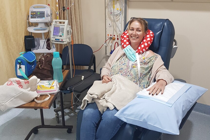 Narelle sitting in a treatment chair smiling, hooked up to machines, wearing neck pillow, arm resting on pillow.