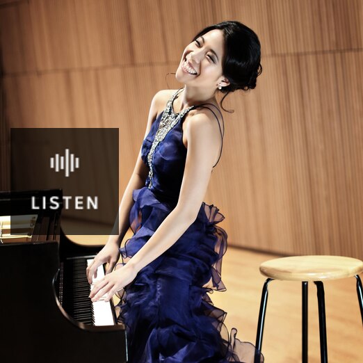 Joyce Yang, wearing a blue dress, stands at the piano with her hands on the keyboard. Has Audio.