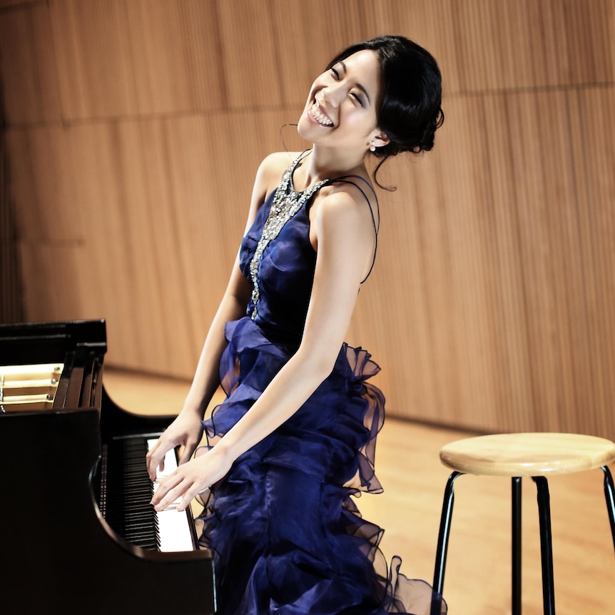 Joyce Yang, wearing a blue dress, stands at the piano with her hands on the keyboard