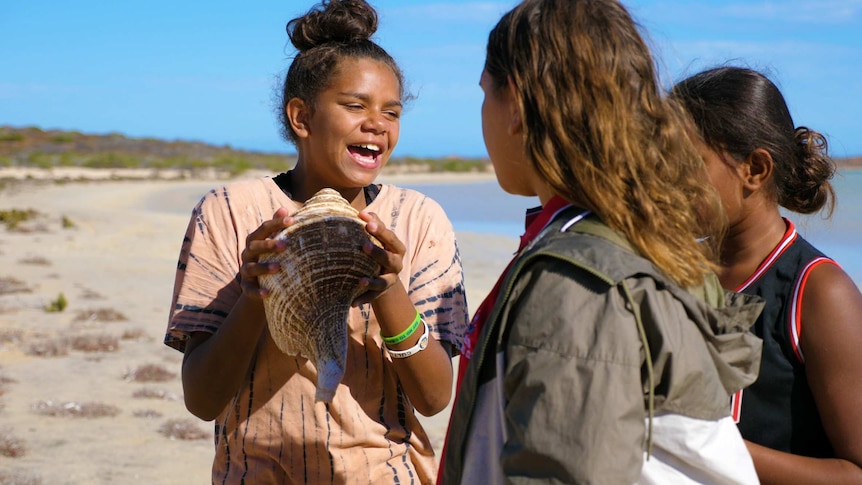 Girls on the beach taking turns blowing into a large shell.