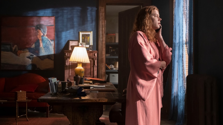 Film still of Amy Adams as Anna dressed in a pink kimono looking out the window, biting her nails in The Woman in the Window