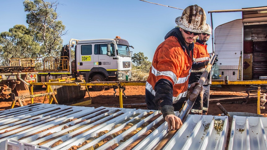 Two men in hard hats and high-vis work clothes place cores of drilled earth into slots, on a tray outside a caravan.