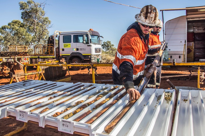 Two men in hard hats and high-vis work clothes place cores of drilled earth into slots, on a tray outside a caravan.