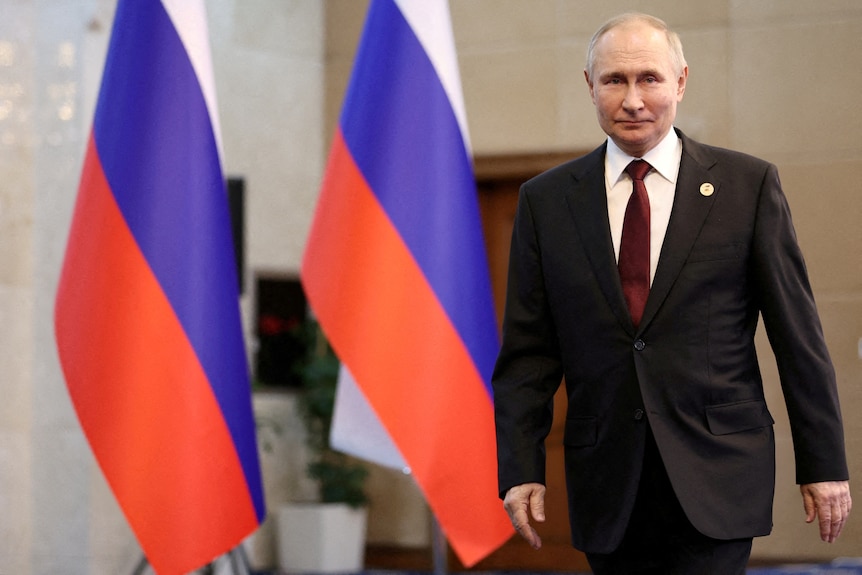 A man in a suit walks past two Russian flags.