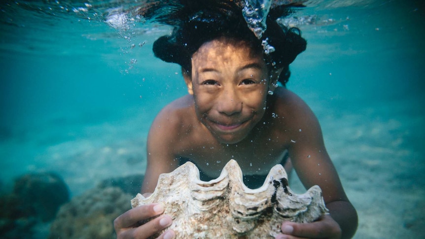 An underwater photograph in clear light blue waters shows a boy holding a closed white clamshell