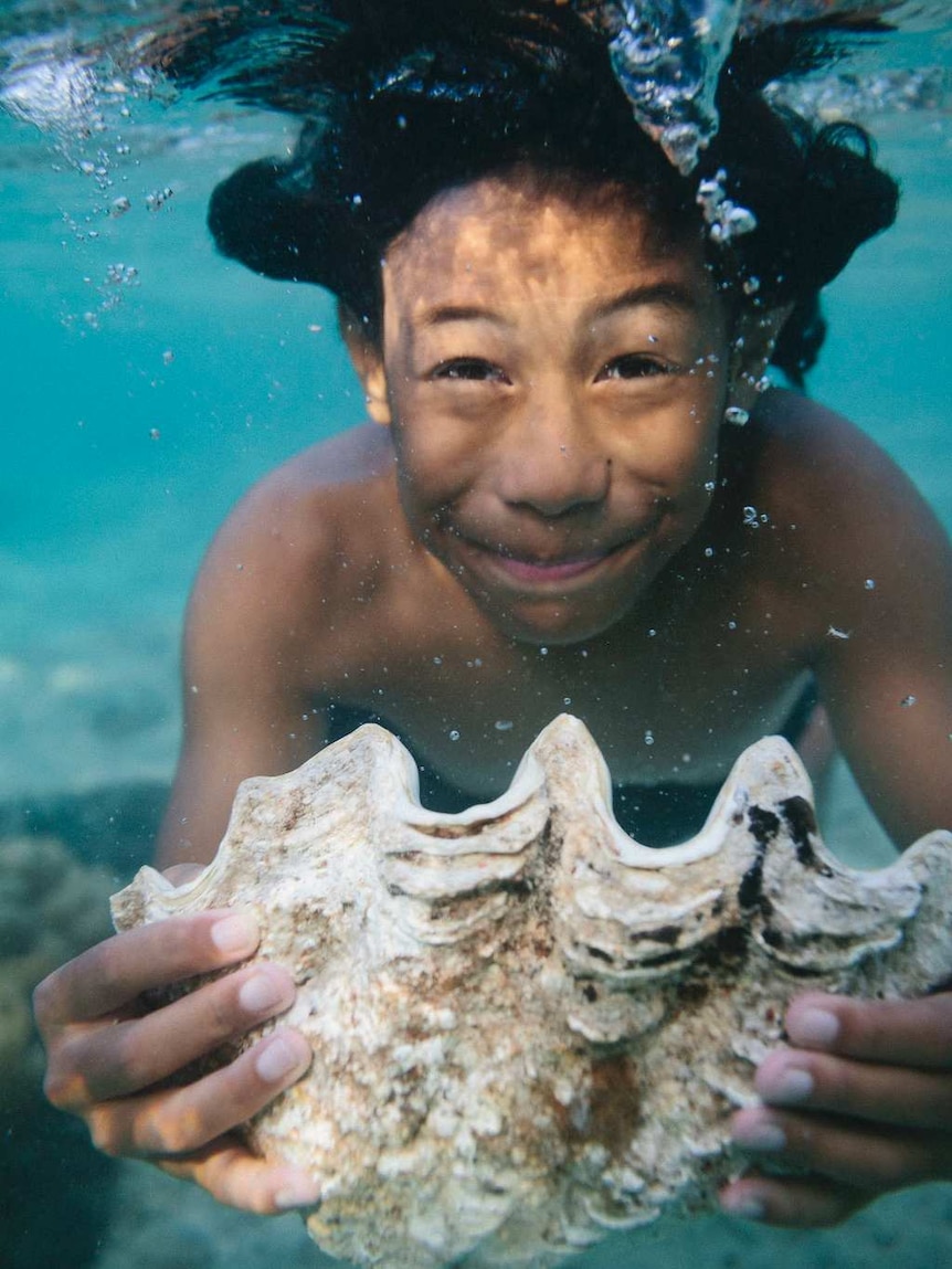 An underwater photograph in clear light blue waters shows a boy holding a closed white clamshell