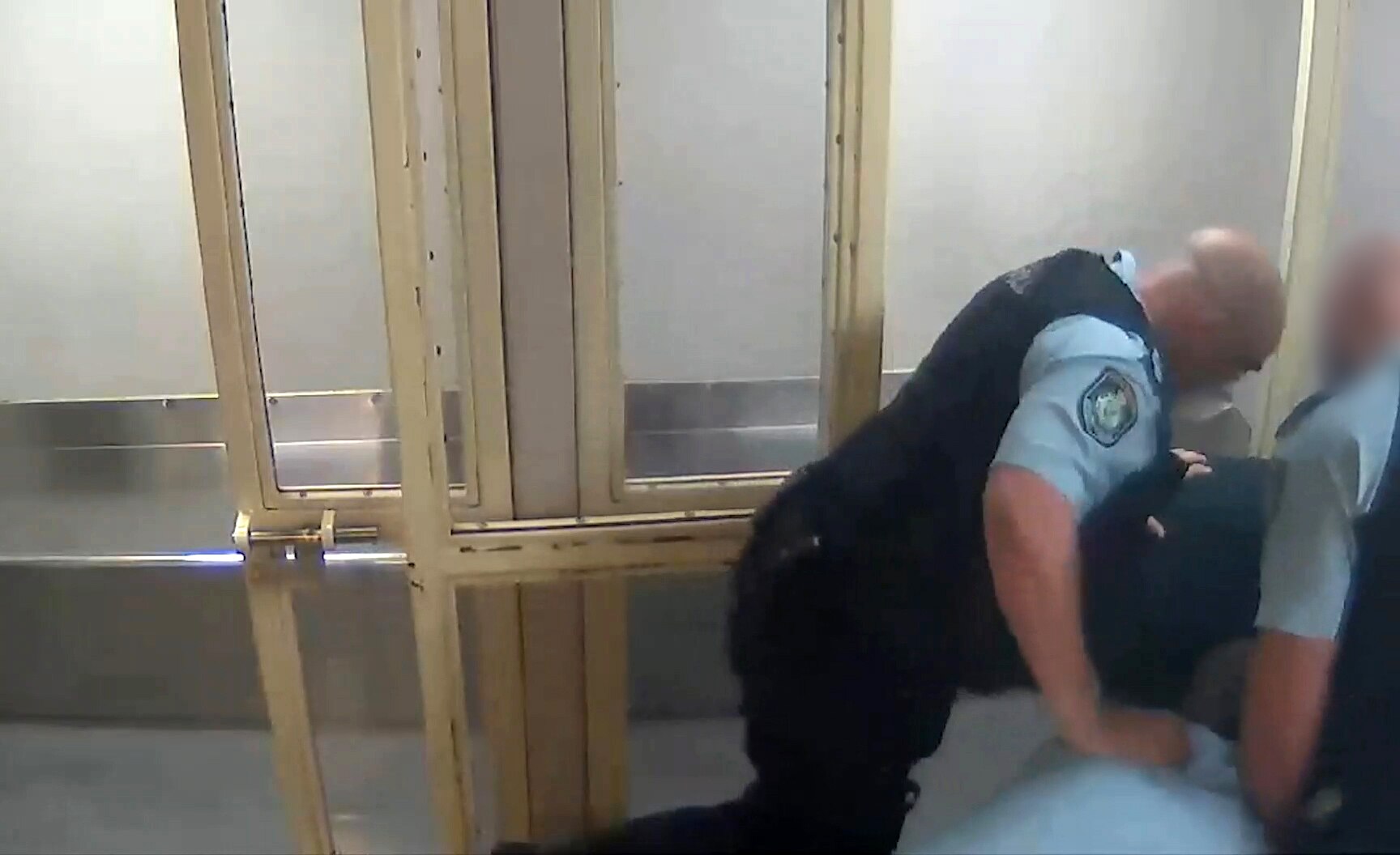 A frame of a cctv video of police cells. Several police officers are crowded over a man as another cop raises his knee. 