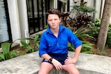 A teenage boy in a blue shirt and shorts sitting in front of a garden.