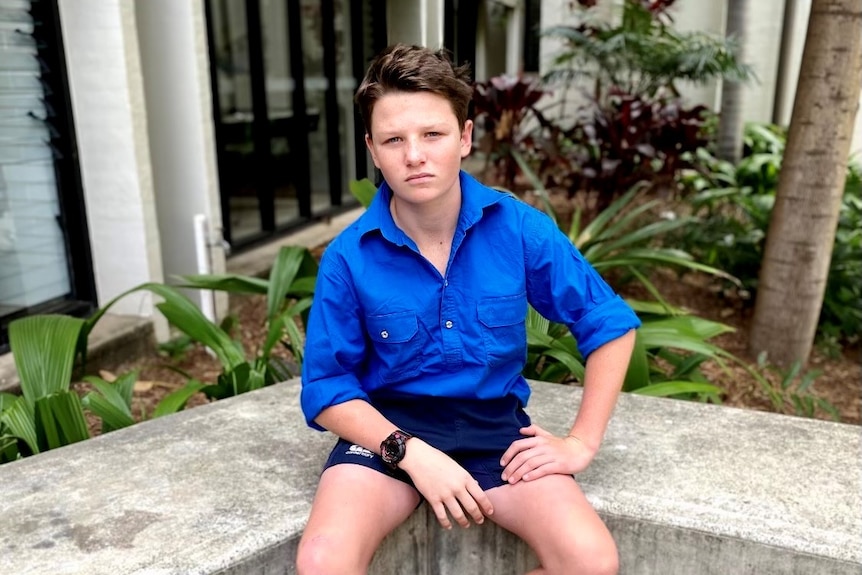 A teenage boy in a blue shirt and shorts sitting in front of a garden.