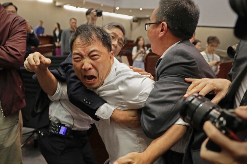 A man with an extremely angry face is held back by security guards inside Hong Kong's parliament.