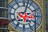 A British Union Jack flag flies in front of the clock face of Big Ben in London with black scaffolding to the left of the image.