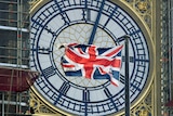 A British Union Jack flag flies in front of the clock face of Big Ben in London with black scaffolding to the left of the image.