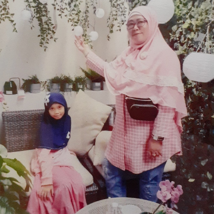 An Indonesian woman in a pink veil stands in a garden, while a small girl in a purple veil sits on a bench
